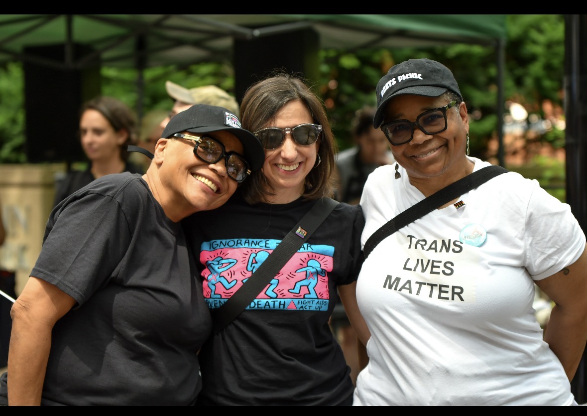 Community members smiling together at an outdoor Pride event, wearing shirts that affirm trans lives and LGBTQ+ liberation